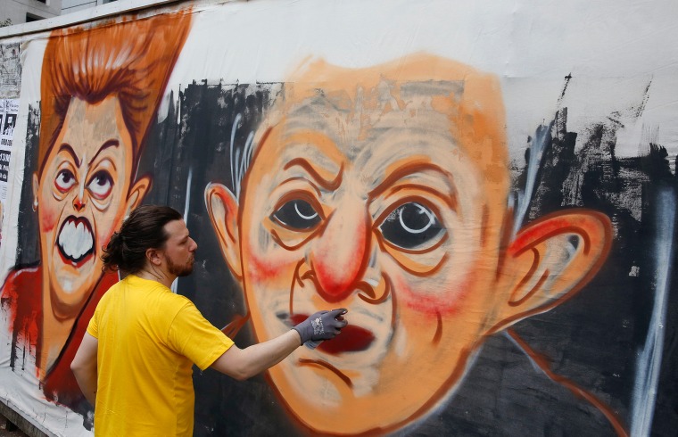 Image: A street artist spray paints caricatures of Brazil's former President Luiz Inacio Lula da Silva, center, and current President Dilma Rousseff