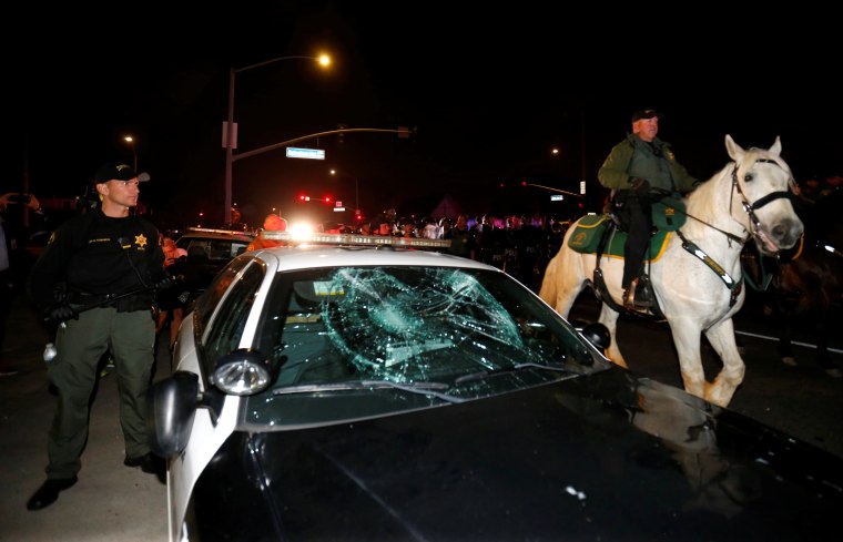 Image: Police on horseback and in riot gear pass by a damaged police car as they break up a demonstration outside Republican U.S. presidential candidate Donald Trump's campaign rally in Costa Mesa