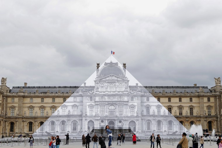 Image: Tourists walk around the JR project at the Louvre Pyramid