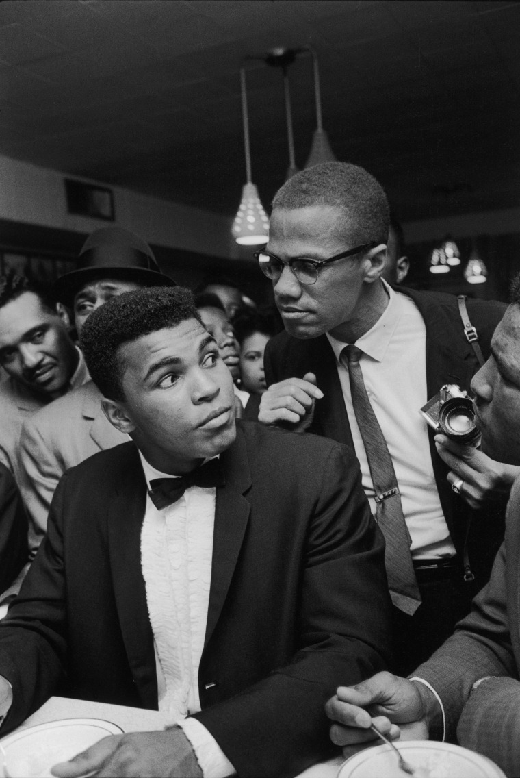 March 1964 - Miami: Black Muslim leader Malcolm X (2R), teasingly leaning on shoulder of tux-clad Cassius Clay (now Muhammad Ali) (L), who is sitting at soda fountain counter, surrounded by jubilant fans after he beat Sonny Liston for the heavyweight championship of the world.
