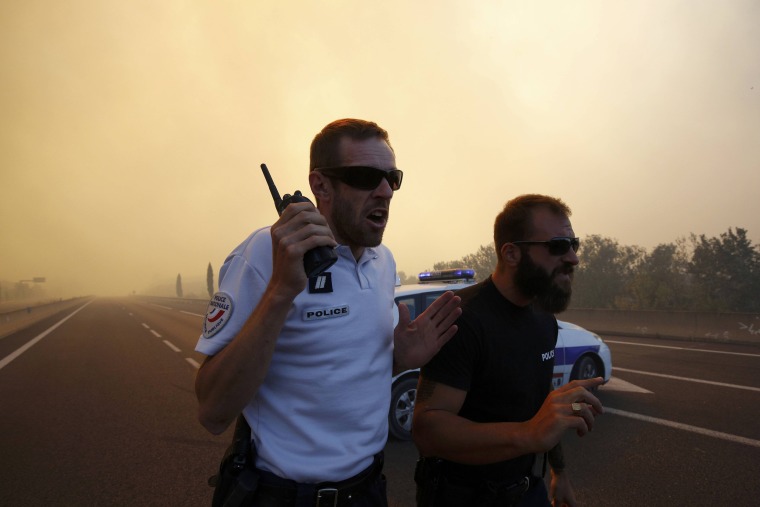 Image: French police stand at a roadblock as smoke fills the sky and blocks the sun as fires burn north of Marseille
