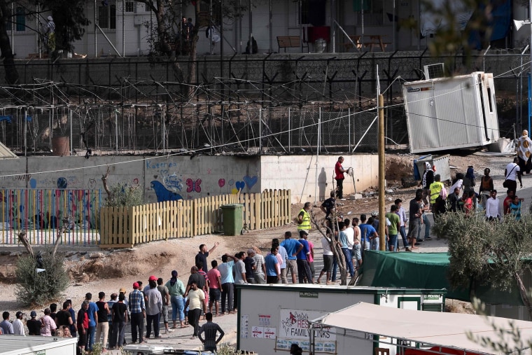 Image: Migrants wait next to burned tents at the camp