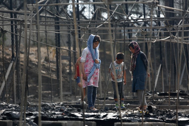 Image: Migrants stand among the remains of a burned tent at the Moria migrant camp, after a fire that ripped through tents and destroyed containers during violence among residents, on the island of Lesbos