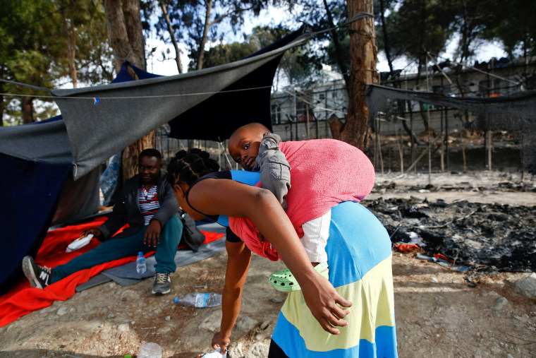 Image: A migrant woman carries a baby as she stands next to the remains of a burned tent at the Moria migrant camp