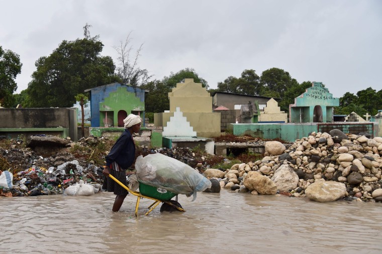 Image: A woman pushes a wheelbarrow while walking in a partially flooded street in Port-au-Prince