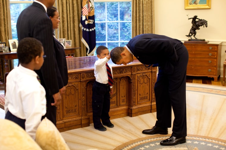Image: President Barack Obama bends over so the son of a White House staff member can pat his head during a visit to the Oval Office