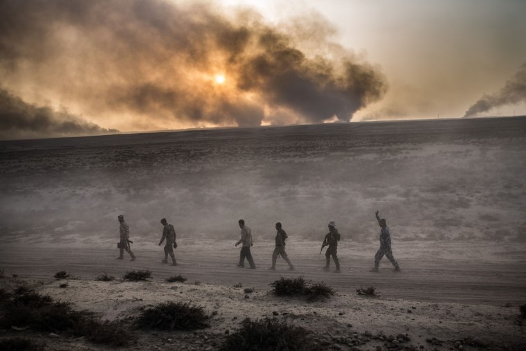 Image: Iraqi soldiers walk on a road as smoke billows from the Qayyarah area