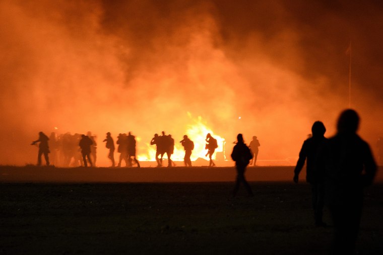 Image: Smoke billows as migrants start fires during overnight clashes ahead of the camp's evacuation.