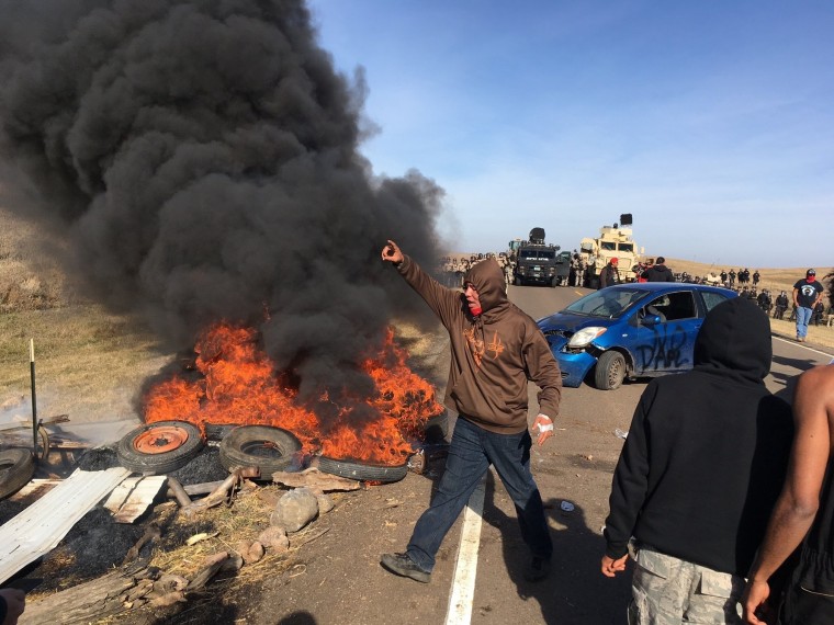 Image: Demonstrators stand next to burning tires as armed soldiers and law enforcement officers assemble