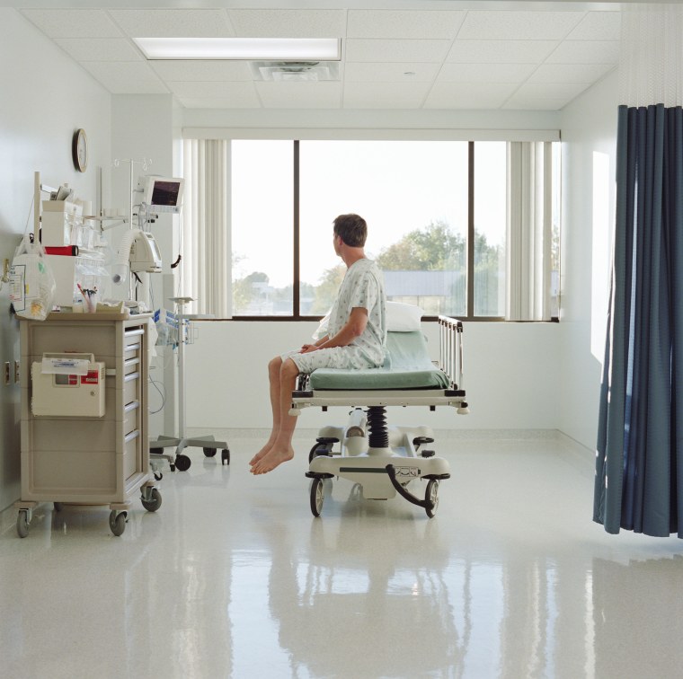 Man in examination gown sitting on bed in hospital room, side view