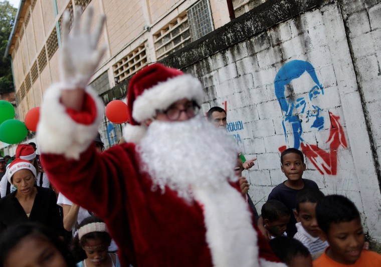 Image: Santa Claus walks past a mural depicting Venezuela's late President Hugo Chavez during a visit to residents of the slum of Petare in Caracas