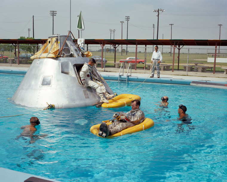 Apollo 1 crew members practice water egress procedures in a swimming pool at Ellington Air Force Base in Houston, Texas. Astronaut Edward H. White II rides on the life raft in the foreground. Roger B. Chaffee sits in the hatch of the boilerplate model of the spacecraft. Virgil I. Grissom waits inside the spacecraft.