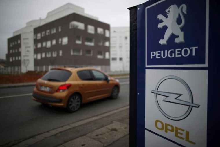 A Peugeot car drives past the logos of French car maker Peugeot and German car maker Opel at a dealership in Villepinte