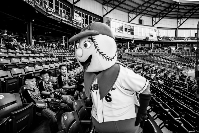 Image: Members of a Cedar Rapids minor league baseball team named the Kernals laugh with a game mascot.
