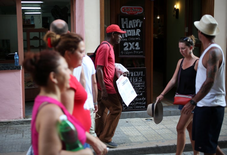 Image: People walk past a bar and a sign advertising "Happy hour" in Havana