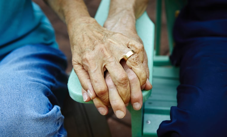 Image: An elderly couple holds hands on a park bench