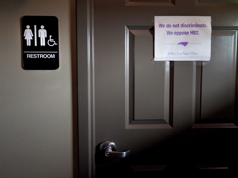 Image: A unisex sign and the "We Are Not This" slogan are outside a bathroom.