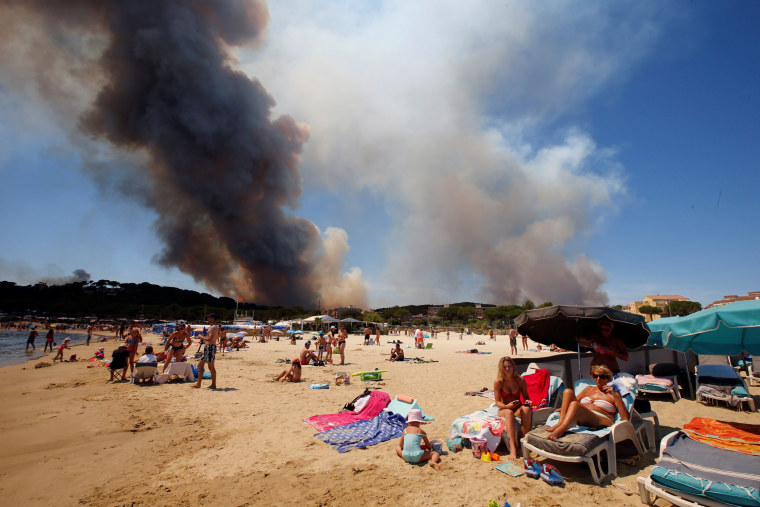 Image: Smoke fills the sky above a burning hillside as tourists relax on the beach in Bormes-les-Mimosas