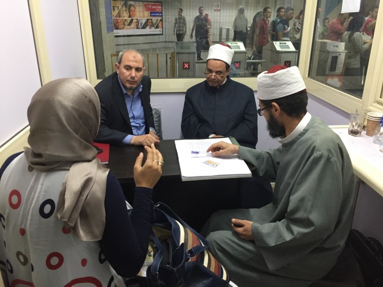 Image: Muslim clerics huddle with passengers at a table in a windowed kiosk