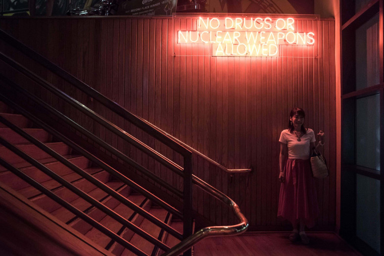 Image: A woman poses for a friend under a sign reading "No drugs or nuclear weapons allowed"