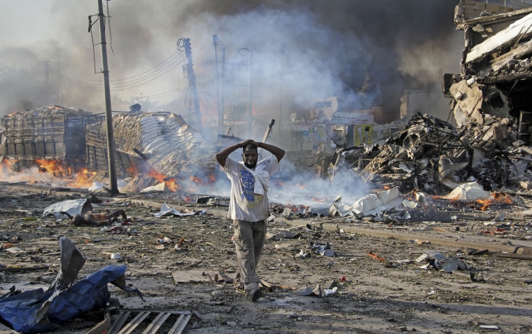 Image: A Somali gestures as he walks past a dead body, left, and destroyed buildings at the scene of a blast