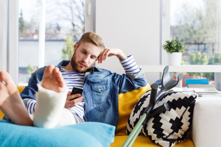 Image: A man sitting on sofa with a broken leg uses a smart phone