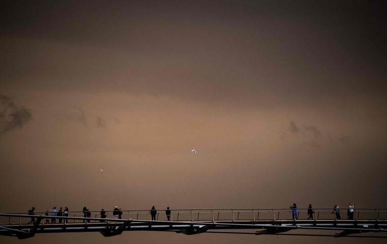 Image: Pedestrians cross the Millennium Footbridge with the sky darkened over London