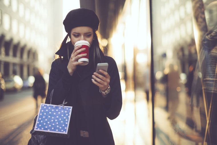 Image: A woman carrying a shopping bag drinks coffee while looking at her mobile phone