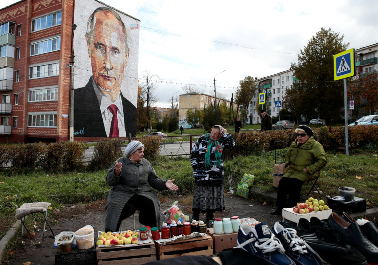 Image: Elderly women wait for customers as they sell their self-made food products at a street market, with a graffiti depicting Russian President Vladimir Putin on the wall of a house seen in the background, in the town of Kashira, outside Moscow