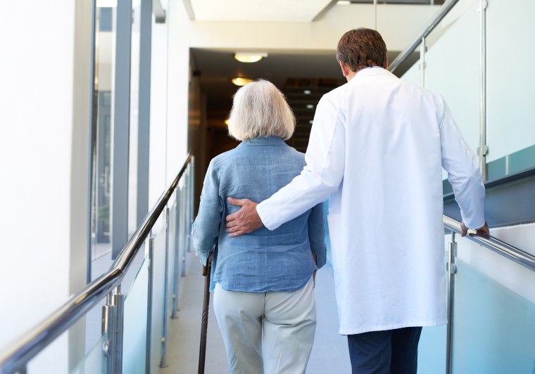 Image: A doctor assists an elderly woman along a hospital corridor
