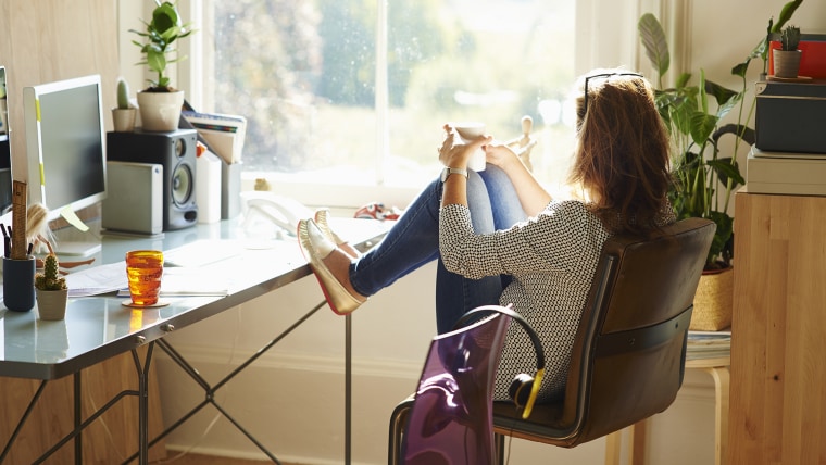 Pensive woman looking through window with feet up on desk in sunny home office