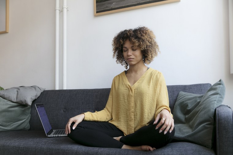 Image: Young woman sitting on couch at home next to laptop meditating