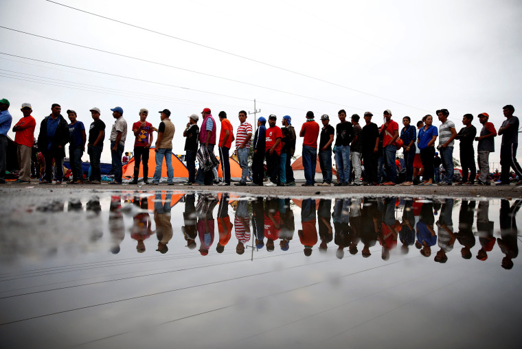 Image: Migrants stand in a line to receive cups of coffee in a makeshift camp at a gas station where the migrants wait for buses which take them to the Mexican border city of Tijuana