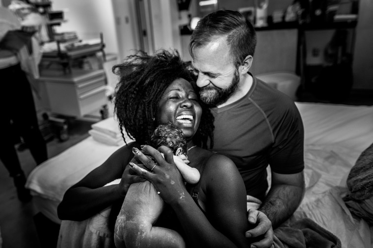 A dad smiling at an exhausted but happy mom, holding their newborn, won third place for black and white photography in the 2019 Birth Becomes Her Image Contest.