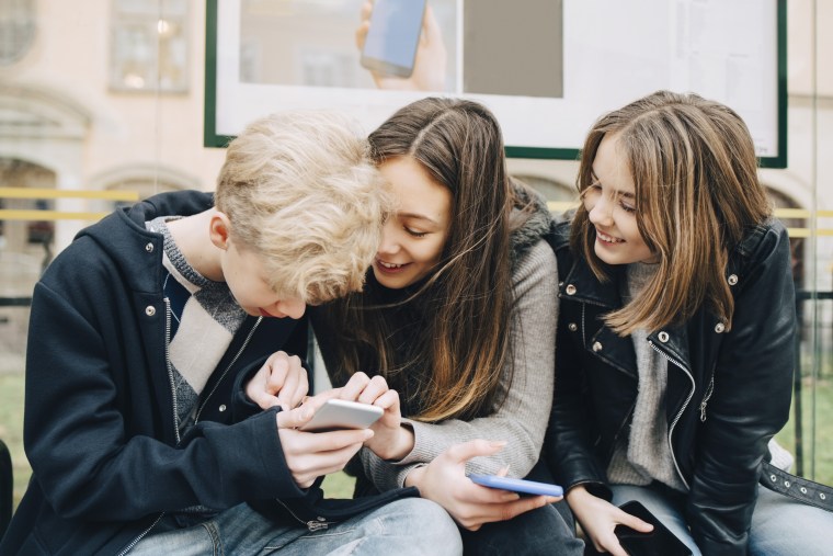Smiling friends using mobile phone while sitting at bus stop