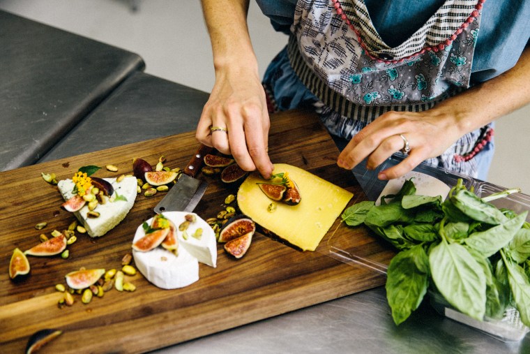 Woman preparing variety cheese plate with figs, nuts, pistachios, basil on wooden chopping board