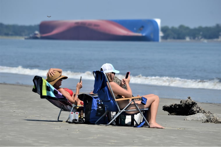 Sun bathers on Jekyll Island's Driftwood Beach look at their phones as the Golden Ray cargo ship sits capsized off the Georgia coast on Sunday.