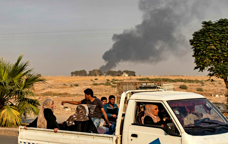 Image: Civilians ride a pickup truck as smoke billows behind them after an attack by Turkey in Syria 