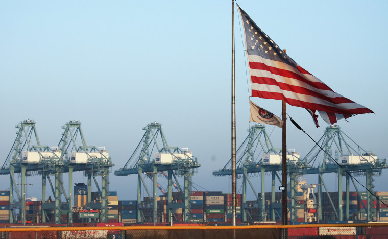 Image: An American flag flies with shipping containers stacked at the Port of Los Angeles in the background, which is the nation's busiest container port
