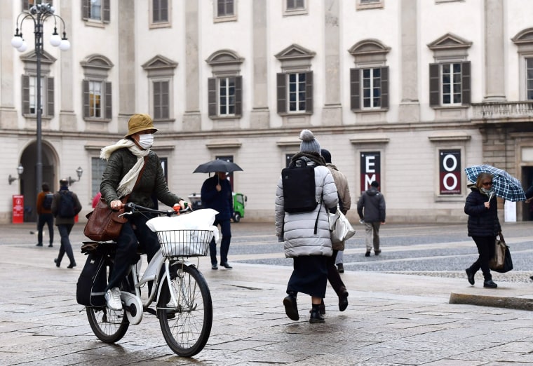 Image: A woman wearing a protective mask crosses Piazza Duomo on a bicycle in Milan