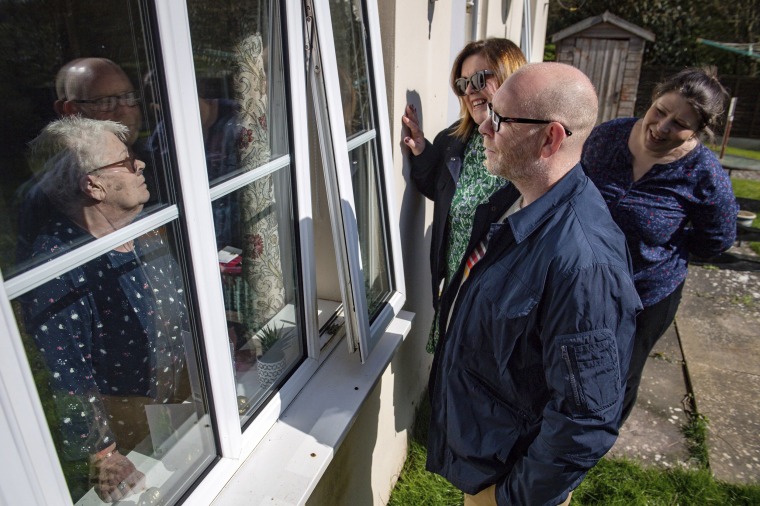 Image: Olive Trotman, left, is visited by her son Mark, his wife, Denise, and his sister, Kelly, for Mother's Day in Napton, England, on March 22, 2020. Olive suffers from a pulmonary disease, and is taking precaution by speaking through a window to limit