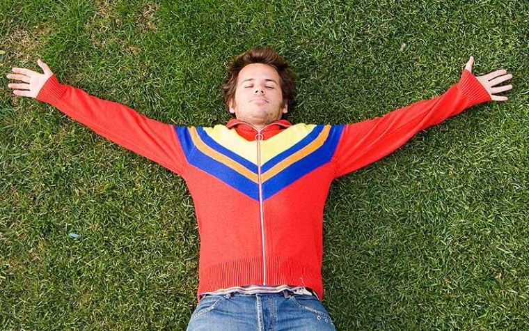 Teenager in red sweater with blue and yellow lies on grass with eyes closed and arms spread