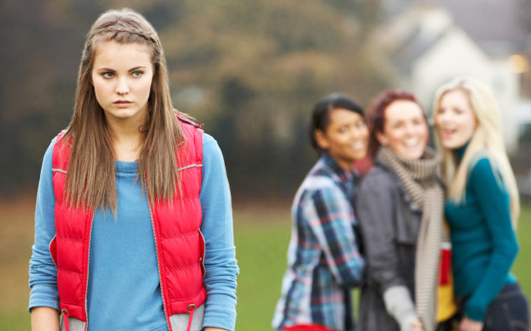 Girl in red vest looking uncomfortable as bullies talk behind her back