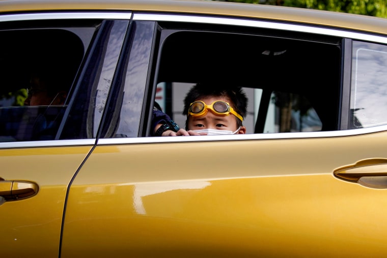 Image: A boy with a face mask and goggles looks out the window of a passing car in Wuhan on April 14, 2020.