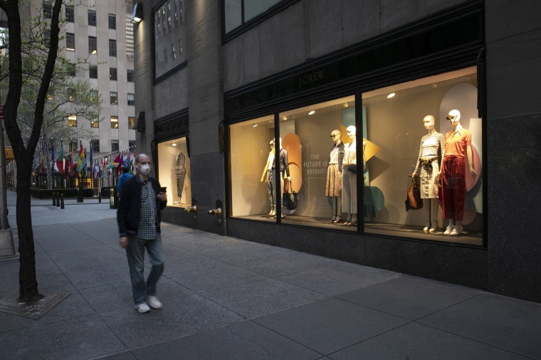 Image: A man wearing a mask walks by a window display at a J Crew store in Rockefeller Center