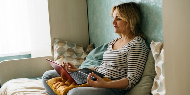 A middle-aged woman sitting in a yoga pose at home on a sofa in casual closes  with laptop and meditating