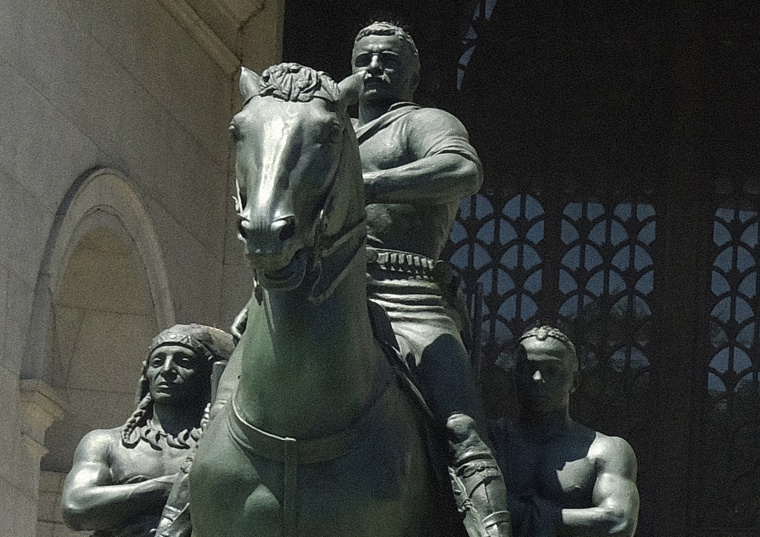 A statue of President Theodore Roosevelt on a horse with an Indigenous person walking alongside him on his right and an African on his left side near the entrance to the Museum of Natural History in New York.