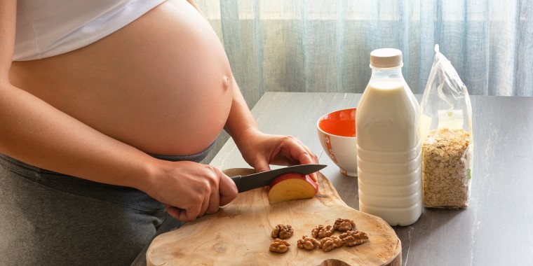 Young and beautiful pregnant woman preparing healthy breakfast at home. Cropped image of pregnant woman cooking a breakfast with oat flakes, apple, milk and walnut.