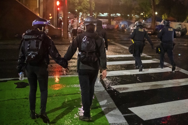 Image: Two protesters hold hands while walking near Portland police officers during an Indigenous Peoples Day of Rage protest