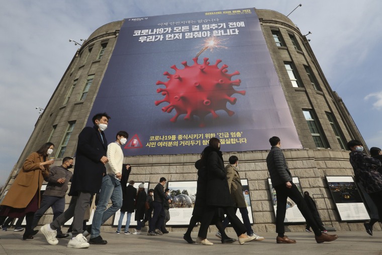 Image: People wearing face masks as a precaution against the coronavirus walk under a banner emphasizing an enhanced social distancing campaign in front of Seoul City Hall in Seoul,